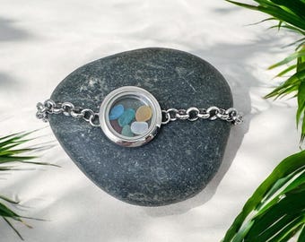May include: A silver bracelet with a round locket filled with colorful stones. The bracelet is resting on a dark gray stone, with green leaves in the background. The locket contains blue, green, yellow, and white stones.
