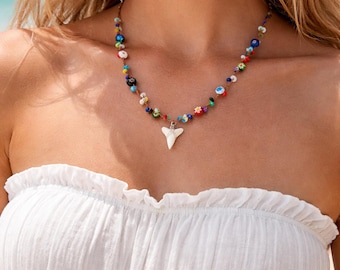 May include: A colorful beaded necklace with a white shark tooth pendant. The necklace features a variety of small, round beads in multiple colors. The shark tooth is white and has a pointed shape.