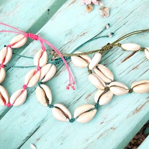 May include: Three seashell bracelets with adjustable cords in pink, green, and teal. The bracelets are made of white cowrie shells strung together. The background is a light blue wooden surface with scattered flower petals.