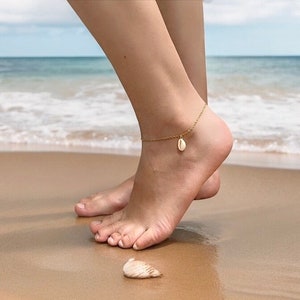 Peut inclure: Un bracelet de cheville doré avec un petit pendentif en forme de coquillage. Le bracelet est porté sur une cheville sur une plage de sable avec l'océan en arrière-plan. Un coquillage repose sur le sable près du pied.