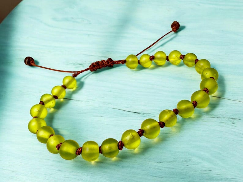 May include: A beaded bracelet featuring translucent, chartreuse-colored beads strung on a brown cord. The bracelet has an adjustable sliding knot closure. The beads are round and evenly spaced, creating a simple, elegant design.