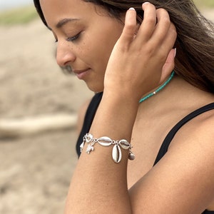 May include: Silver bracelet with large, oval seashells and small charms. The bracelet is worn on an arm, with a turquoise beaded necklace in the background. The bracelet is a beach-themed accessory, suitable for summer.