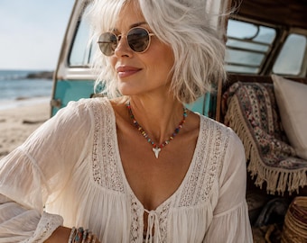 May include: A white long-sleeved blouse with lace detailing and a front tie closure, paired with a necklace featuring a white shark tooth pendant on a strand of small, multi-colored beads, and round-framed sunglasses with dark lenses.