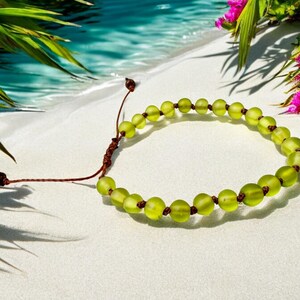 May include: A beaded bracelet with light green, round beads and brown string. The bracelet is on a white surface with a tropical beach background, including palm leaves and water.