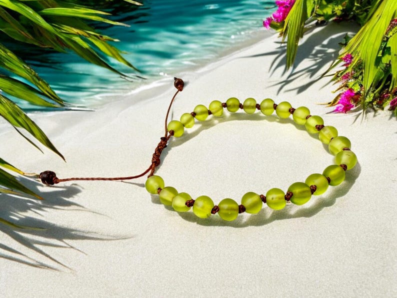 May include: A beaded bracelet with light green, round beads and brown string. The bracelet is on a white surface with a tropical beach background, including palm leaves and water.