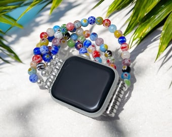 May include: A silver smartwatch with a black screen is paired with a beaded watch band. The band features colorful glass beads in various colors, including blue, red, yellow, and green. The watch and band are displayed on a white surface with green leaves.