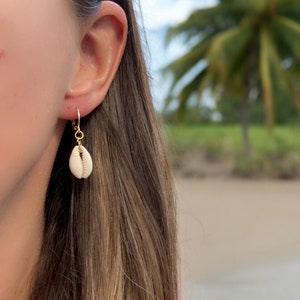 Peut inclure: Boucles d'oreilles dorées avec un petit pendentif coquillage de couleur crème. Les boucles d'oreilles sont portées sur une oreille, avec de longs cheveux brun clair visibles. L'arrière-plan montre une scène de plage avec des palmiers et un haut rose.