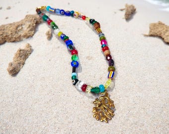 May include: A colorful beaded necklace with a gold-colored coral pendant rests on a sandy surface. The necklace features a variety of glass beads in red, blue, green, yellow, and other colors. The pendant is a detailed, stylized coral design.