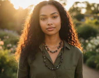 May include: A woman wearing a green collared shirt and two beaded necklaces. The necklaces have dark green and black beads. The woman has long, dark curly hair. The background is a sunlit garden with green foliage.