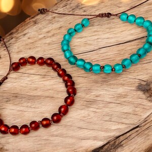 May include: Two beaded bracelets. One is a deep red color, and the other is a bright turquoise. Both bracelets are made of round beads strung on a brown cord. The bracelets are displayed on a wooden surface.