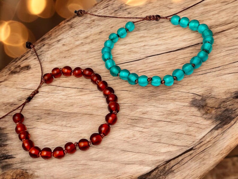 May include: Two beaded bracelets. One is a deep red color, and the other is a bright turquoise. Both bracelets are made of round beads strung on a brown cord. The bracelets are displayed on a wooden surface.