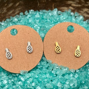 May include: Two pairs of pineapple stud earrings, one silver and one gold, presented on brown circular cards. The earrings are set against a backdrop of light blue, crushed glass-like material.
