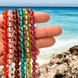 May include: A hand displays six strands of colorful beaded bracelets. The beads are in shades of red, white, brown, teal, yellow-green, and pink. The background shows a beach with turquoise water and a blue sky.