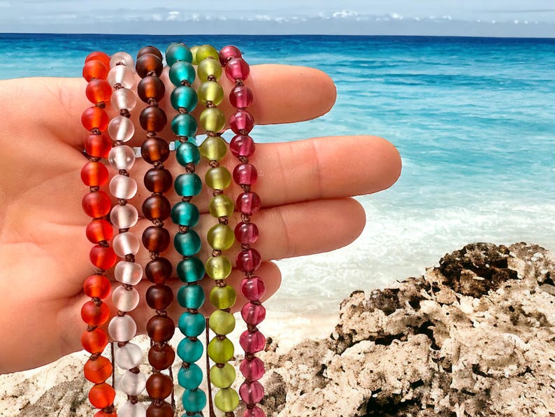 May include: A hand displays six strands of colorful beaded bracelets. The beads are in shades of red, white, brown, teal, yellow-green, and pink. The background shows a beach with turquoise water and a blue sky.