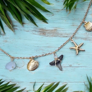 May include: A gold chain necklace featuring ocean-themed charms. The charms include a seashell, starfish, shark tooth, and sea glass. The necklace is presented on a light blue wooden surface, decorated with green palm leaves.