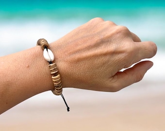 May include: A beach-themed bracelet featuring a white seashell and wooden beads. The bracelet is strung on a black cord and is worn on a wrist. The background shows a blurred view of the ocean.