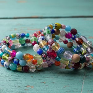 May include: Three colourful beaded bracelets with a variety of shapes and sizes. The beads are in shades of blue, pink, green, yellow, and clear, with some featuring patterns. The bracelets are stacked on a turquoise surface.