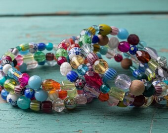 May include: Three colorful beaded bracelets with a variety of shapes and sizes. The beads are in shades of blue, pink, green, yellow, and clear, with some featuring patterns. The bracelets are stacked on a turquoise surface.