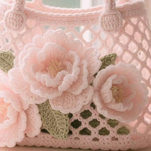 May include: A light pink crochet handbag with a lattice design. The bag features three large, three-dimensional crochet flowers in a matching pink, with green leaves. The handles are also crocheted.