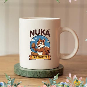 May include: White ceramic mug with the word "NUKA" in brown letters. The mug features a cartoon squirrel holding a nut, with the words "NUTT BUTTER" below. The mug is on a green coaster, with a floral border.