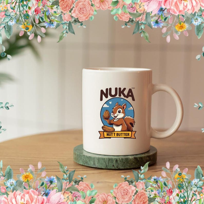 May include: White ceramic mug with the word "NUKA" in brown letters. The mug features a cartoon squirrel holding a nut, with the words "NUTT BUTTER" below. The mug is on a green coaster, with a floral border.