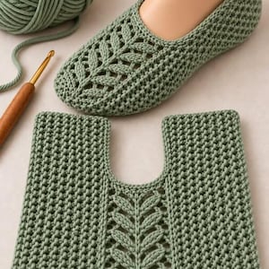 May include: A sage green crocheted slipper with a textured leaf pattern on the top, accompanied by a matching square crocheted piece featuring a central leaf design, a ball of sage green yarn, and a wooden crochet hook.