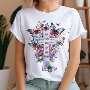May include: White t-shirt with a colorful cross design featuring butterflies and flowers. The cross has a prayer written on it that reads "Our Father who art in Heaven, hallowed be thy name. Thy kingdom come. Thy will be done, on earth as it is in Heaven. Give us this day our daily bread. And forgive us our trespasses, as we forgive those who trespass against us. And lead us not into temptation, but deliver us from evil. For thine is the kingdom, and the power, and the glory, forever and ever. Amen."