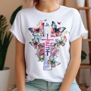 May include: White t-shirt with a watercolor design of a cross surrounded by butterflies and flowers. The word "FAITH" is written vertically on the cross.