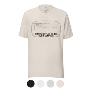 May include: A light beige t-shirt with a humorous design. The design features a text box with code and the phrase "CONFUSED? YEAH, ME TOO. BUT IT COMPILES." The shirt has a crew neck and short sleeves.