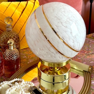 May include: A white glass globe lamp with gold accents and a white glitter finish. The lamp is on a gold base and sits on a mirrored surface.