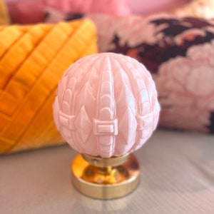 May include: A pink glass lamp with a decorative, geometric pattern. The lamp has a gold base and sits on a white surface.