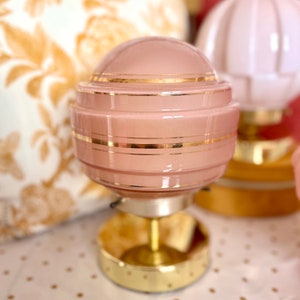 May include: A pink glass lampshade with gold trim sits on a gold metal base. The lampshade has a round shape and is decorated with horizontal gold stripes.