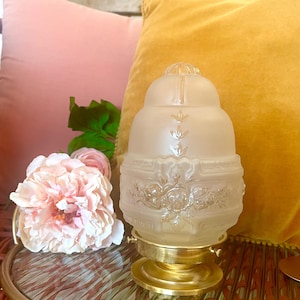 May include: A vintage-style frosted glass lamp shade with floral and leaf detailing. The shade is mounted on a brass base. The background includes pink and yellow pillows and a pink flower arrangement, creating a soft, romantic aesthetic.