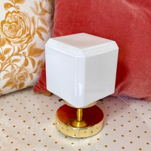 May include: A white cube-shaped lamp with a brass base. The lamp is sitting on a white surface with gold polka dots.