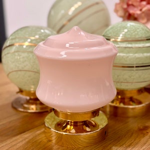 May include: A pink glass lamp with a gold base and accents. The lamp has a rounded, flared shape with a pointed top. Other green glass lamps with gold accents are in the background. The lamps are on a wooden surface.