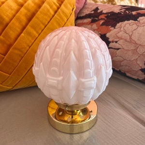 May include: A pink glass lampshade with a geometric pattern sits on a gold metal base. The lampshade has a textured surface and is shaped like a sphere.