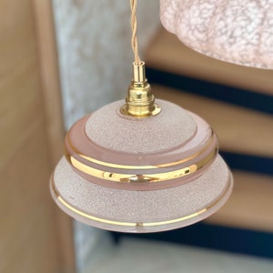 May include: A pink glass pendant light with a gold trim and a textured surface. The light is hanging from a gold cord.