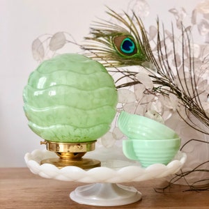 May include: A green glass lampshade with a wavy pattern sits on a white pedestal cake stand.  The lampshade is on a brass base.  Three green glass cups are stacked next to the lampshade.