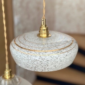 May include: A white glass pendant light with gold accents. The glass has a speckled texture and the gold accents are in the form of thin lines.