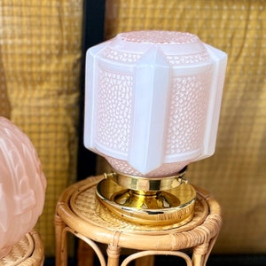 May include: A pink glass lampshade with a textured surface sits on a woven wicker table. The lampshade has a gold metal base.
