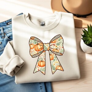 Peut inclure: Sweat-shirt crème avec un grand motif de nœud orné de citrouilles, de feuilles et de motifs floraux orange, vert et marron. Le nœud est bordé de marron foncé. Le sweat-shirt est plié sur une surface en bois clair, à côté d'un jean bleu.