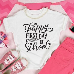 Op de afbeelding: Wit T-shirt met zwarte tekst die "...happy... first day of school" luidt met een potlood-afbeelding en hartjes. Het shirt ligt op een roze en wit chevron-patroon met roze jeans, roze glitter sneakers, een roze hoofdband met een paarse strik en roze zonnebrillen.