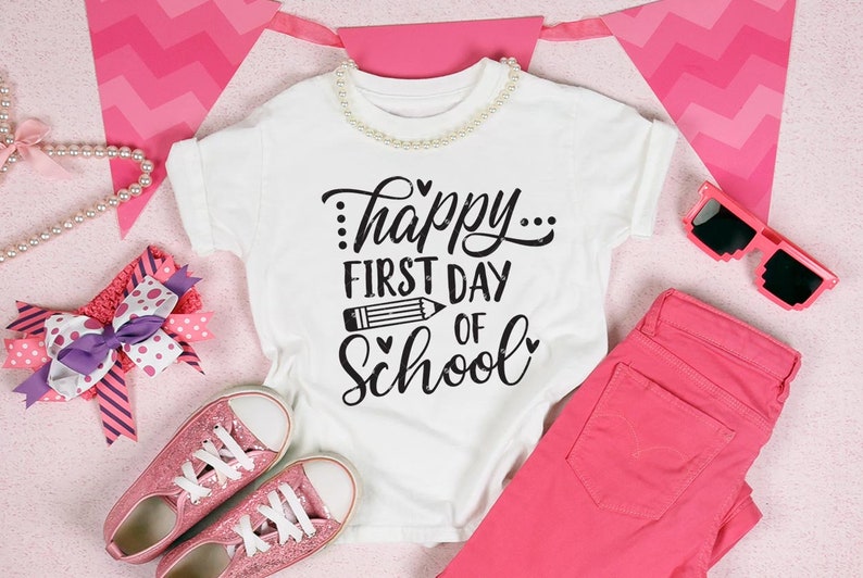 Op de afbeelding: Wit T-shirt met zwarte tekst die "...happy... first day of school" luidt met een potlood-afbeelding en hartjes. Het shirt ligt op een roze en wit chevron-patroon met roze jeans, roze glitter sneakers, een roze hoofdband met een paarse strik en roze zonnebrillen.