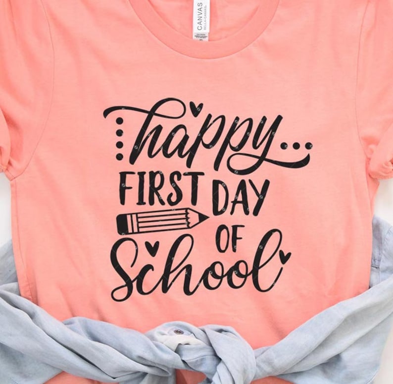 Op de afbeelding: Een perzikkleurig T-shirt met de tekst "...happy... first day of school" in zwarte letters. Een zwart potlood is getekend tussen de woorden "first day" en "of school".