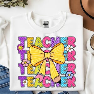 Peut inclure: Sweat-shirt blanc avec le mot "TEACHER" en lettres colorées à pois. Un grand nœud jaune et un dessin de crayon sont centrés, avec des fleurs. Un cadeau idéal pour les enseignants.