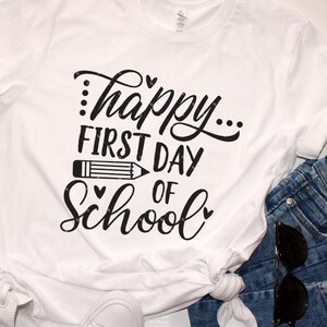 Op de afbeelding: Wit T-shirt met zwarte tekst die "...happy... first day of school" luidt, met een potloodgrafiek en harten.