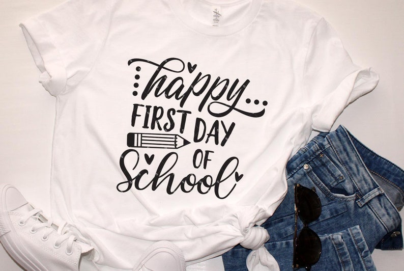 Op de afbeelding: Wit T-shirt met zwarte tekst die "...happy... first day of school" luidt, met een potloodgrafiek en harten.