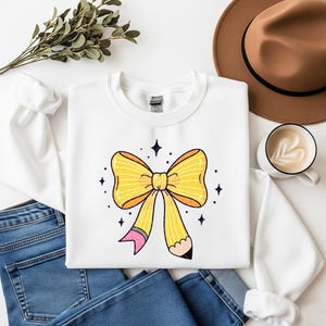 May include: White sweatshirt featuring a large yellow bow design with a pencil detail. The bow has orange accents and is surrounded by small star shapes. The sweatshirt is paired with blue jeans, a brown hat, and a coffee cup.