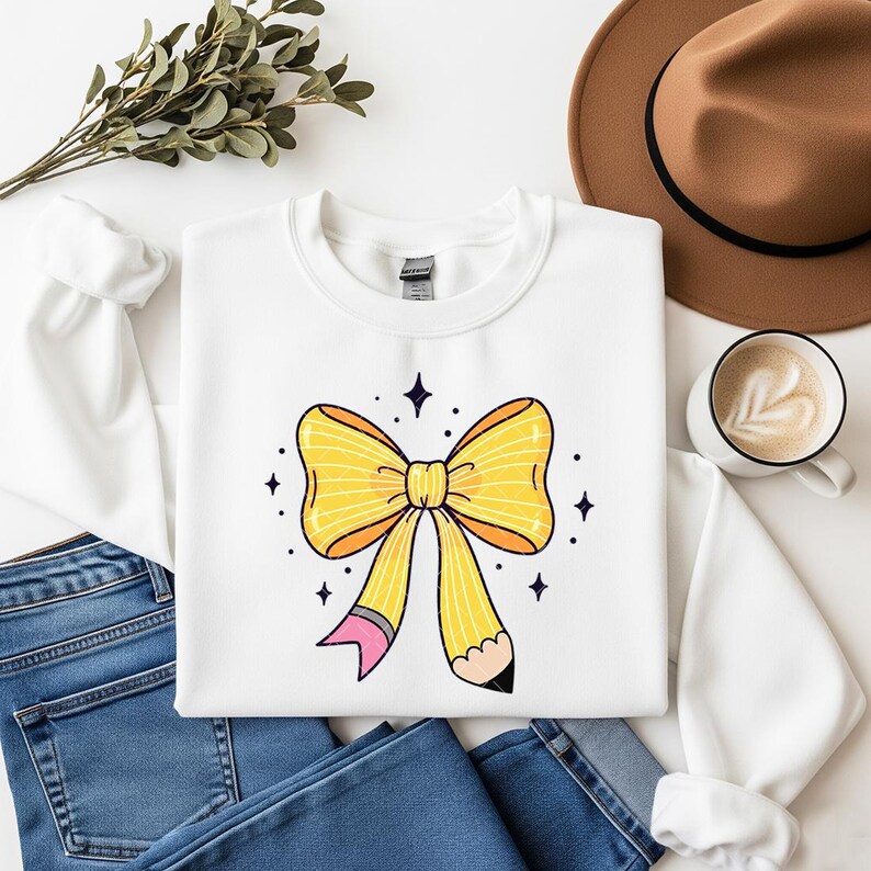 May include: White sweatshirt featuring a large yellow bow design with a pencil detail. The bow has orange accents and is surrounded by small star shapes. The sweatshirt is paired with blue jeans, a brown hat, and a coffee cup.