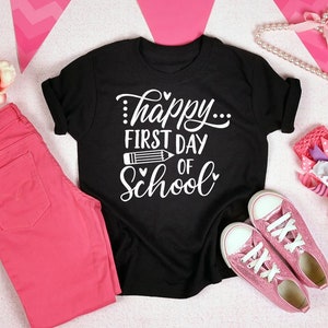 Op de afbeelding: Zwart T-shirt met witte tekst die "...happy... first day of school" zegt met een potlood-afbeelding. Het shirt ligt op een roze oppervlak met roze jeans, een roze hoofdband en een parelketting.